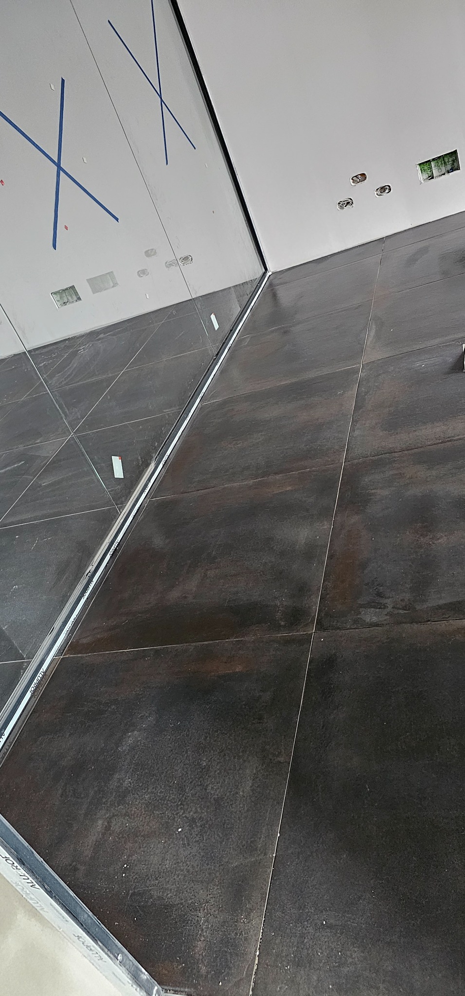 Ciemne, matowe płytki ceramiczne na ścianie, częściowo ułożone, z widocznymi aluminiowymi profilami krawędziowymi i fragmentem sufitu z instalacją elektryczną.