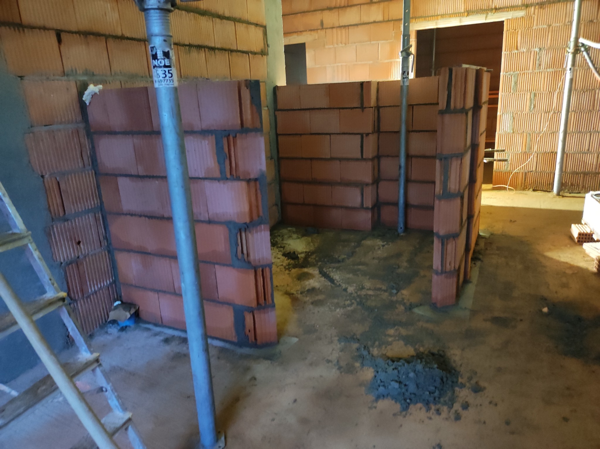 Wznoszenie ścianek działowych z czerwonej cegły ceramicznej, widoczne podpory stalowe i resztki zaprawy na podłodze w surowym wnętrzu.