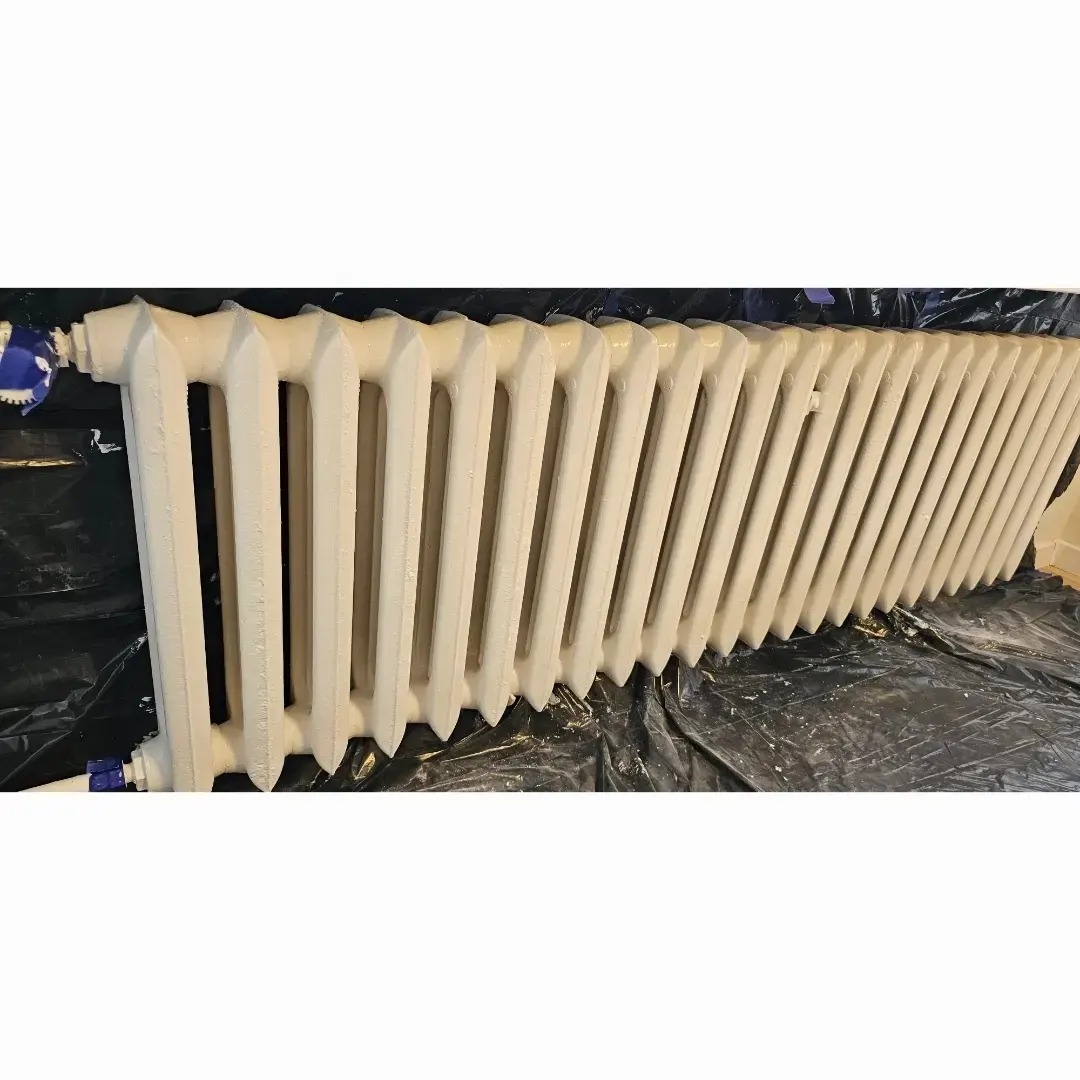 Żeliwny grzejnik pomalowany na biało, zabezpieczony folią malarską, z widocznymi świeżymi śladami farby.