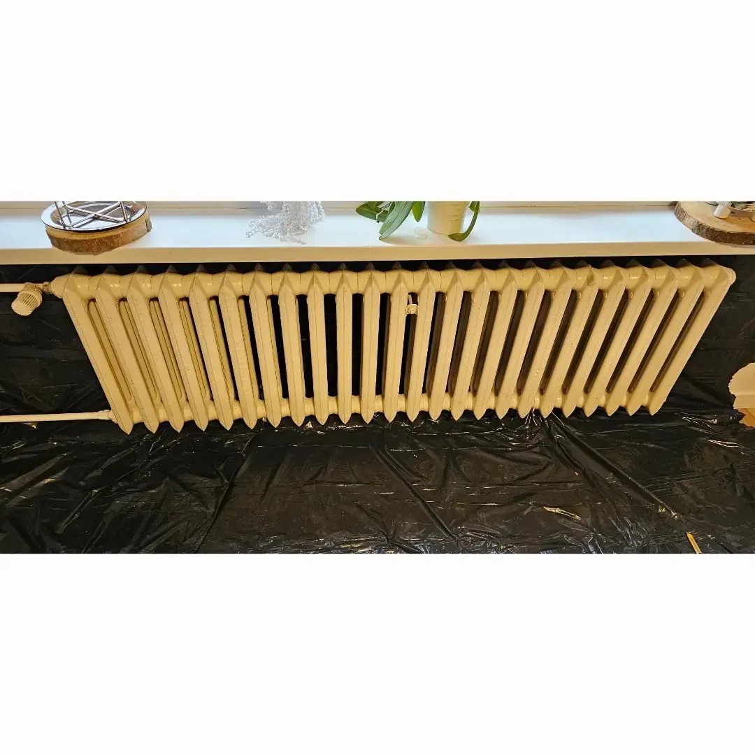 Żeliwny grzejnik, zabezpieczony czarną folią malarską, przygotowany do renowacji, widok z przodu, parapet z dekoracjami w tle.