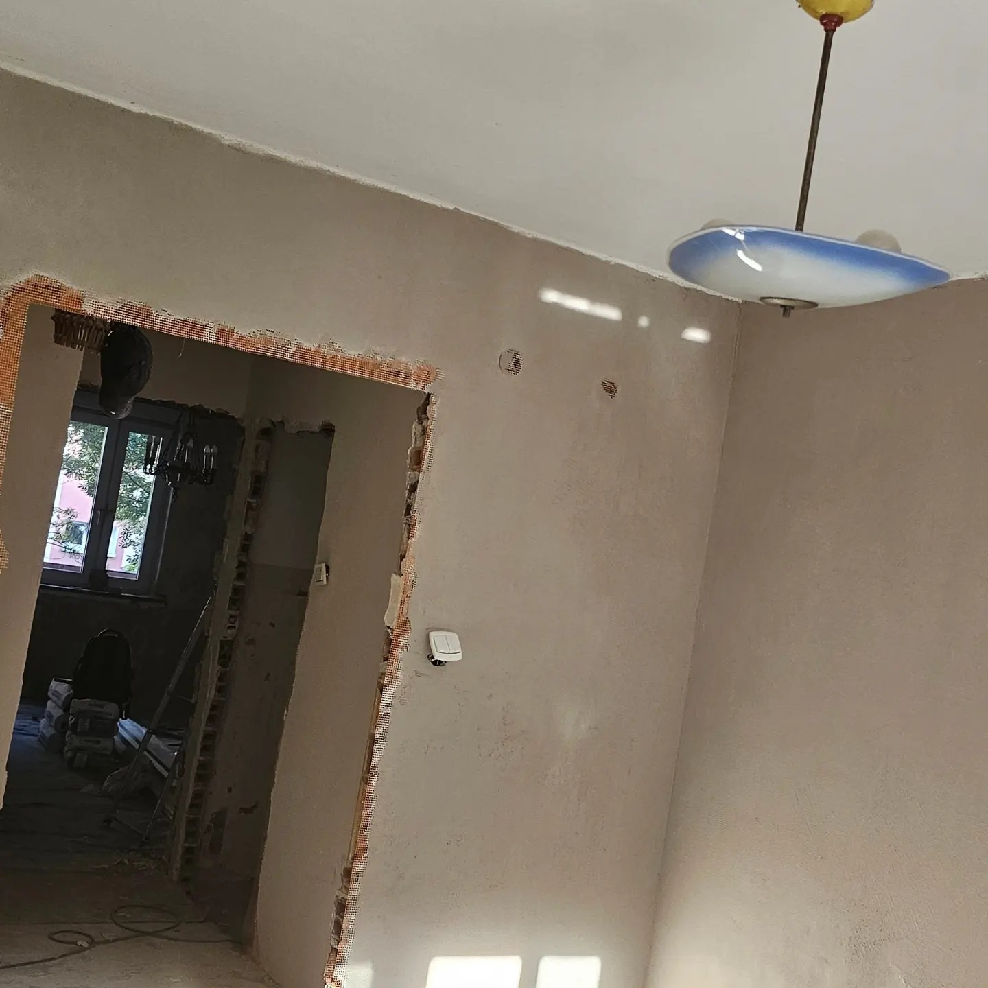 Pokój w trakcie remontu, ściany pokryte świeżo nałożoną warstwą gładzi, widoczne siatki wzmacniające na krawędziach ościeżnicy, w tle worki z materiałami budowlanymi.