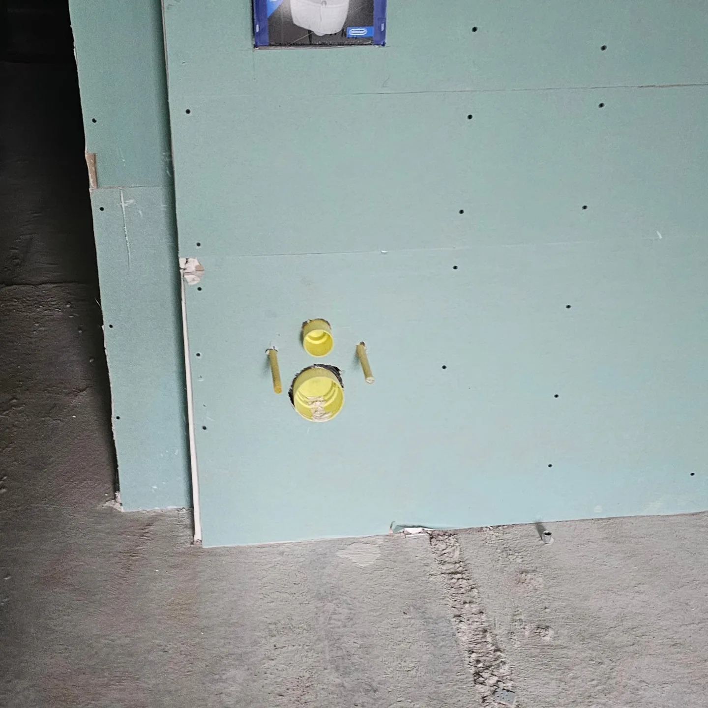 Niebieska płyta karton gips z żółtymi rurami instalacyjnymi wystającymi ze ściany, widoczne ślady po wkrętach i niedoskonałości w wykończeniu.