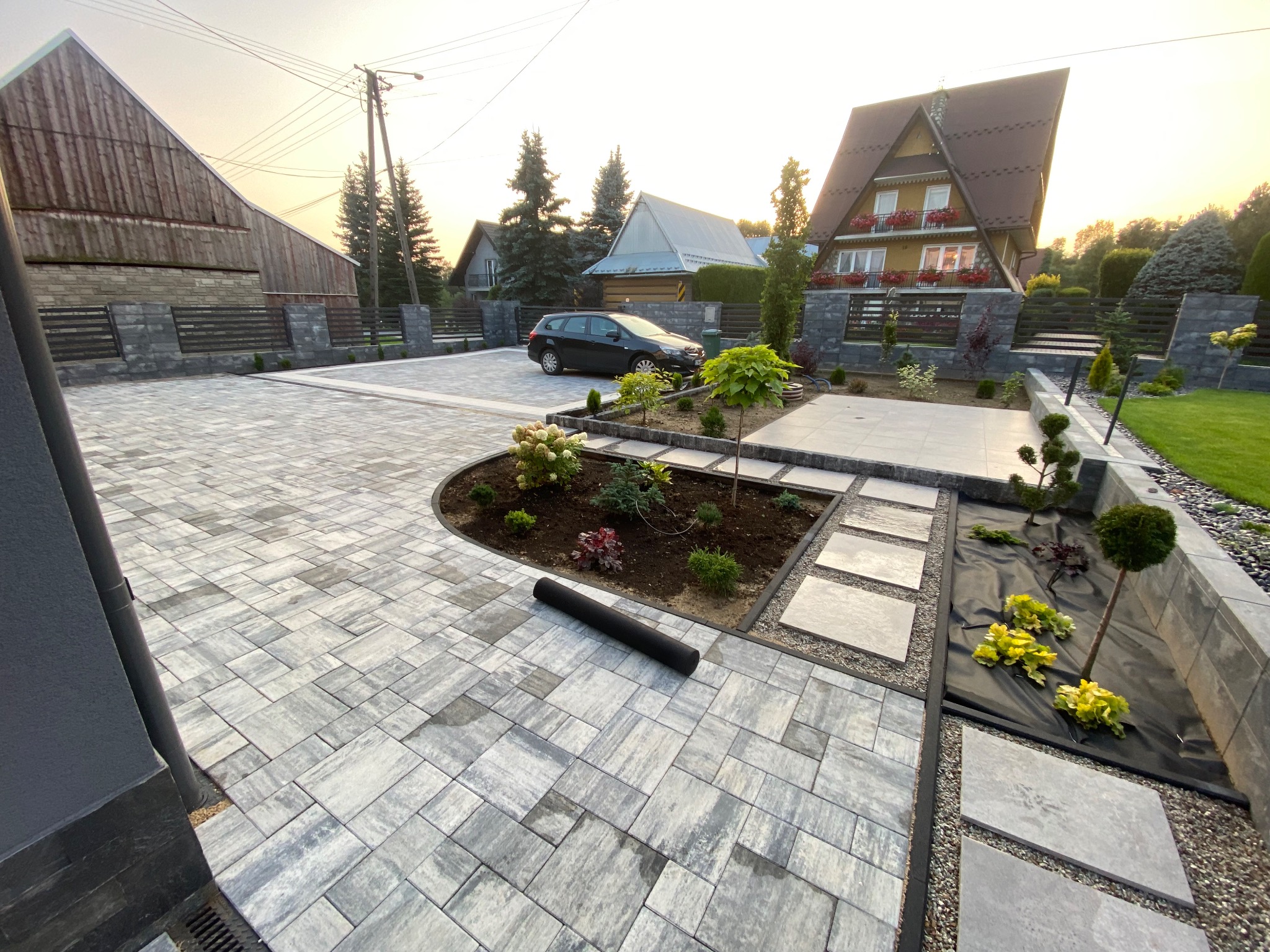 Ułożona szara kostka brukowa na podjeździe i ścieżkach wokół domu z ogrodem, widoczny czarny samochód i fragment domu z czerwonymi kwiatami w oknach.