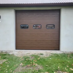 Brązowa segmentowa brama garażowa z trzema okienkami, zamontowana w jasnym budynku z metalowym dachem, widok z zewnątrz na trawnik z opadłymi liśćmi.