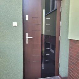 Nowoczesne drzwi wejściowe w kolorze ciemnego drewna z przeszkleniem, zamontowane w budynku z zieloną elewacją i brązową cegłą.