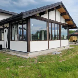 Nowoczesna przeszklona weranda z drewnianą konstrukcją, widok z zewnątrz na trawnik i parasole ogrodowe.