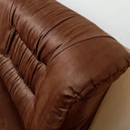 Fragment brązowej, pikowanej kanapy z wyraźną fakturą skóry ekologicznej, oświetlony z lewej strony, ukazujący detale szwów i załamań materiału.