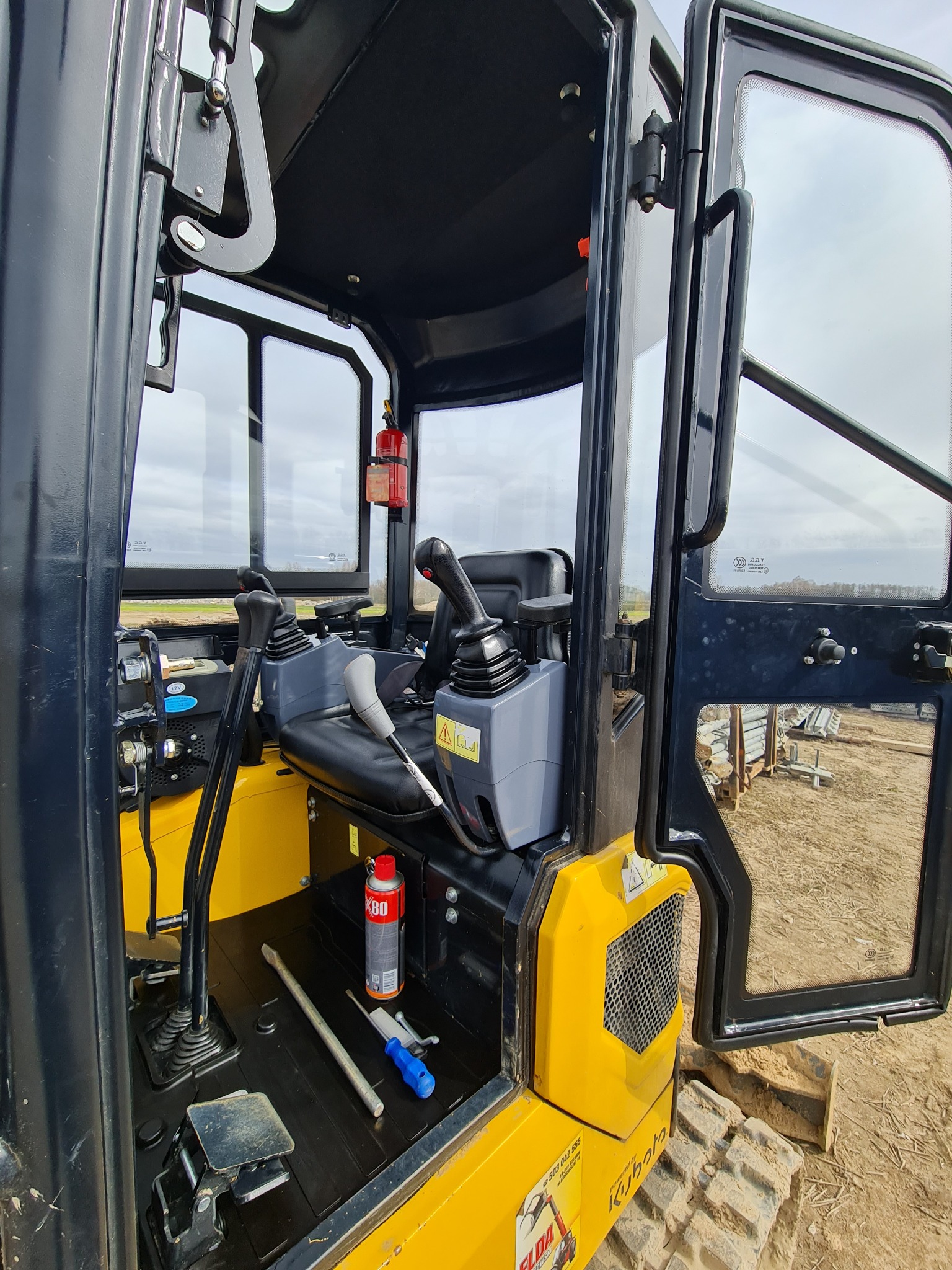 Wnętrze kabiny żółtej minikoparki Kubota z widocznymi dźwigniami sterującymi, fotelem operatora, gaśnicą i narzędziami na podłodze kabiny. Otwarta szyba kabiny z widokiem na plac budowy.