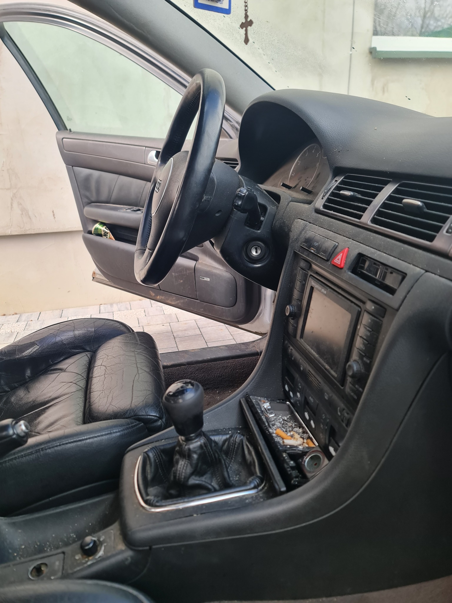 Wnętrze zaniedbanego samochodu Audi A6 C5 z widoczną warstwą kurzu na desce rozdzielczej, popielniczka pełna niedopałków, otwarte drzwi kierowcy i zużyte skórzane fotele.