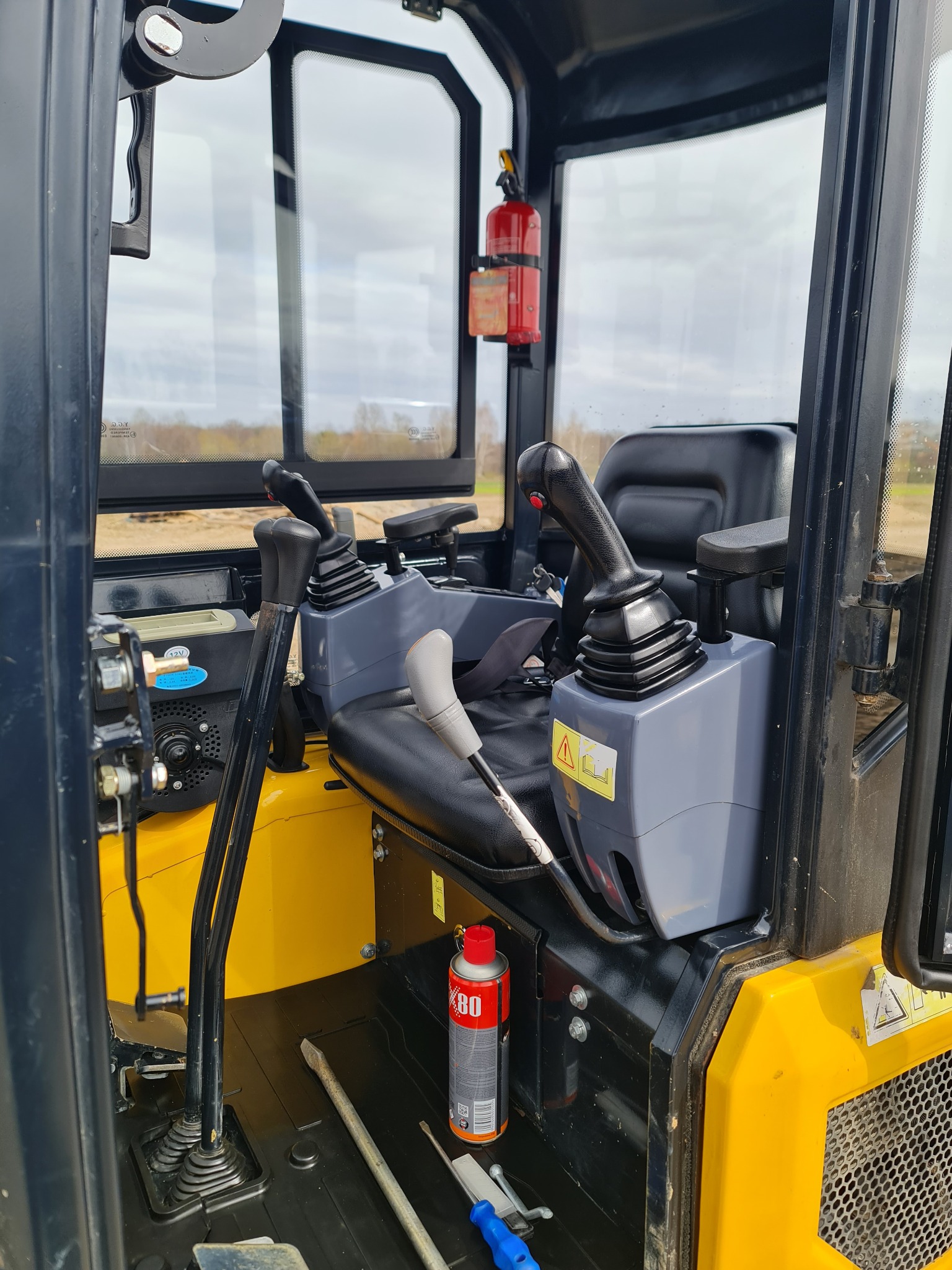 Wnętrze kabiny małej koparki z widocznym fotelem operatora, joystickami sterującymi, gaśnicą przymocowaną do ramy oraz puszką smaru na podłodze.