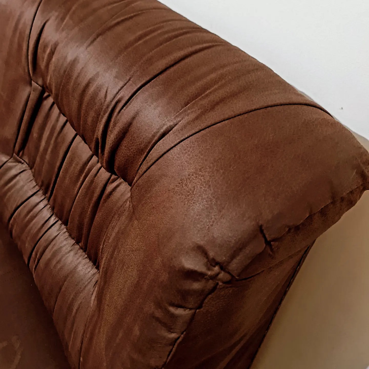 Fragment brązowej, pikowanej kanapy z wyraźną fakturą skóry ekologicznej, oświetlony z lewej strony, ukazujący detale szwów i załamań materiału.