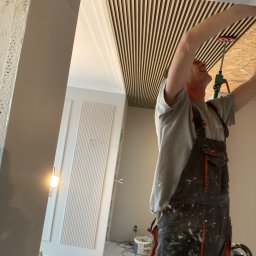 Omilian Tomasz - Montaż drewnianych paneli akustycznych na suficie przez uśmiechniętego mężczyznę w roboczym kombinezonie, widoczne narzędzia i częściowo wykończona ściana z dekoracyjnymi listwami.