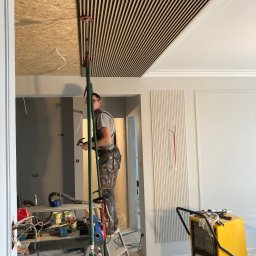 Omilian Tomasz - Montaż drewnianych paneli sufitowych przez montera na drabinie; widoczny sufit z wykończonym fragmentem, narzędzia, agregat malarski i przygotowane ściany w szarym kolorze.