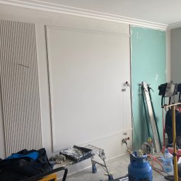 Omilian Tomasz - Wnętrze w trakcie prac wykończeniowych: ściany z dekoracyjnymi panelami i listwami, fragment ściany z płytą gipsowo-kartonową, pracownik z narzędziem, butla gazowa i akcesoria malarskie na podłodze.