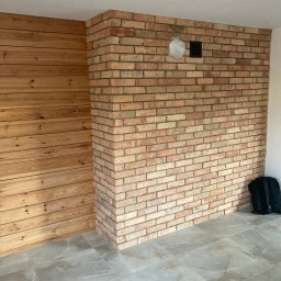 Omilian Tomasz - Fragment pokoju z wykończoną ścianą z cegły klinkierowej, drewnianą boazerią i szarą podłogą z płytek imitujących marmur. W tle widoczny czarny plecak.