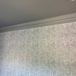 Omilian Tomasz - Fragment pokoju z szarą ścianą pokrytą wzorzystą tapetą w odcieniach szarości i bieli, widoczna listwa przysufitowa i fragment futryny drzwi.