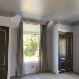Omilian Tomasz - Jasne wnętrze pokoju po remoncie, z dwuskrzydłowymi, ciemnymi szafami wnękowymi z białymi obramowaniami, oknami z kremowymi zasłonami i grzejnikiem żeberkowym pod oknem, wykończone sztukaterią.