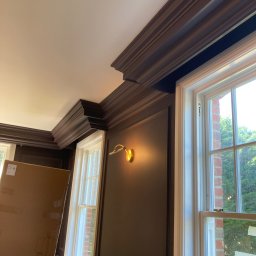 Omilian Tomasz - Eleganckie, ciemnobrązowe boazerie i listwy przysufitowe w pokoju z oknami, widoczna drabinka malarska i zabezpieczone okno folią malarską.