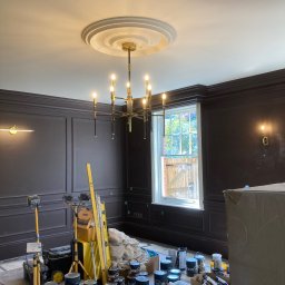 Omilian Tomasz - Pokój po malowaniu z ciemnymi ścianami z panelami, widoczne puszki z farbą, drabina i sprzęt oświetleniowy, żyrandol w stylu retro