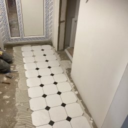 Omilian Tomasz - Układanie białych, ośmiokątnych płytek podłogowych z czarnymi wstawkami w pomieszczeniu z oknem PCV, widoczne narzędzia i warstwa kleju.