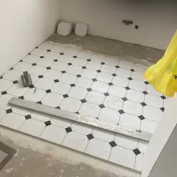 Układanie białych, ośmiokątnych płytek podłogowych z czarnymi kwadratami w narożach, widoczne narzędzia glazurnicze: poziomica, kielnia i ołówek