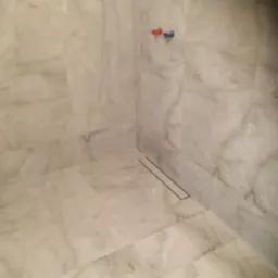 Wykończony brodzik kabiny prysznicowej z odpływem liniowym, wyłożony płytkami imitującymi marmur, widoczne przyłącza ciepłej i zimnej wody.