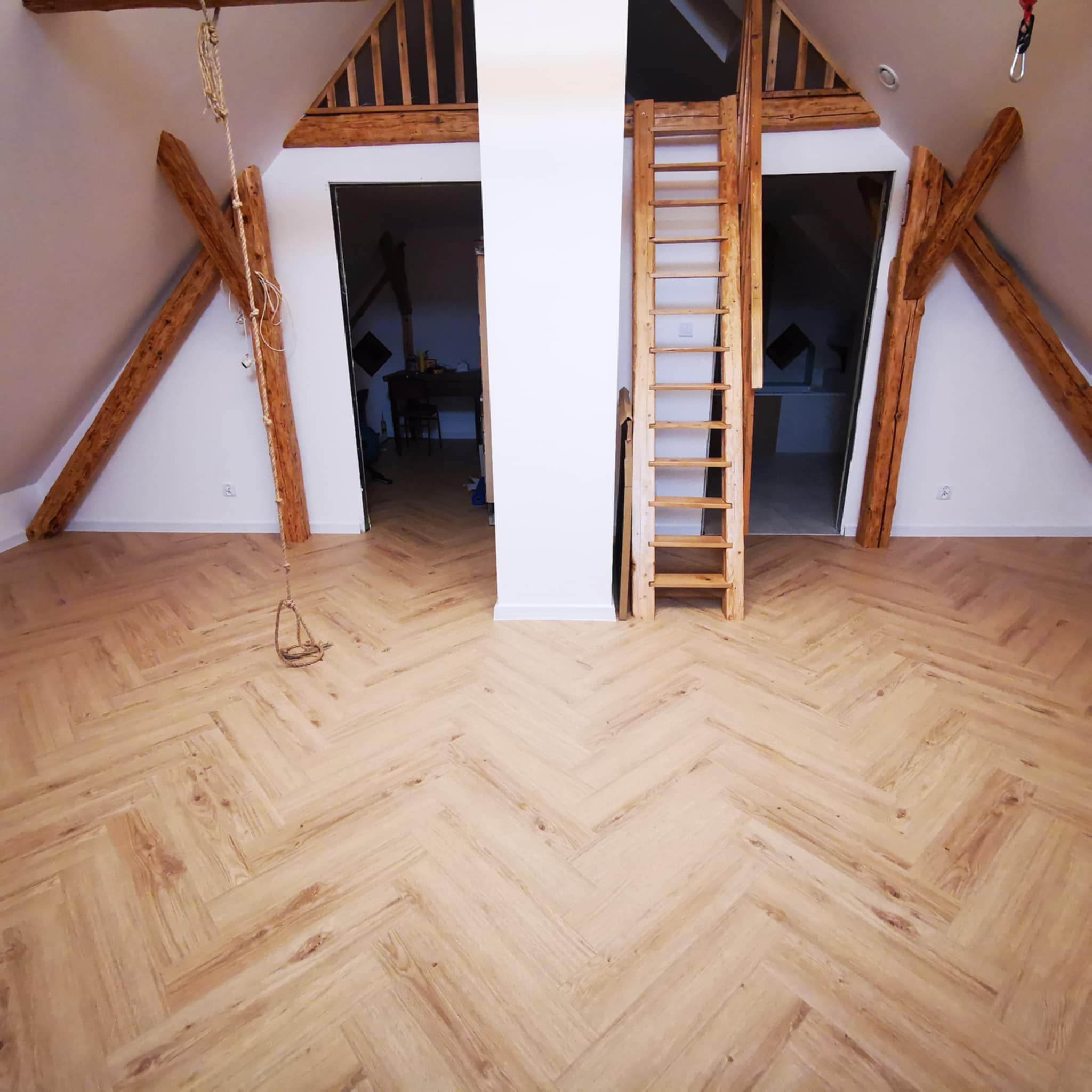 Jasne panele ułożone we wzór jodełki w przestronnym pokoju na poddaszu z drewnianymi belkami stropowymi i drabiną.