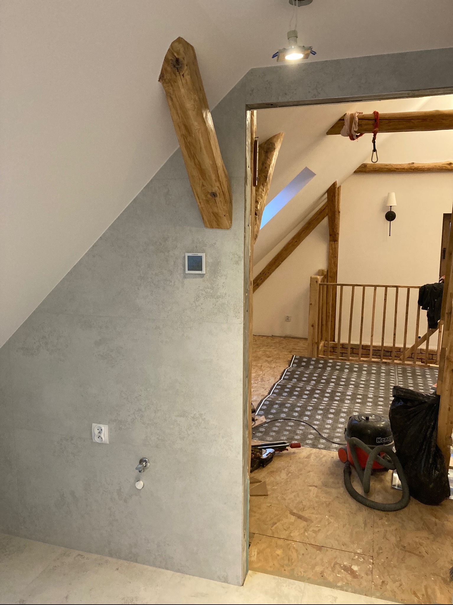 Poddasze w trakcie prac wykończeniowych z widoczną konstrukcją drewnianą, oknem dachowym i przygotowaną podłogą z izolacją, przez próg widoczny odkurzacz przemysłowy i ściana wykończona szarą...