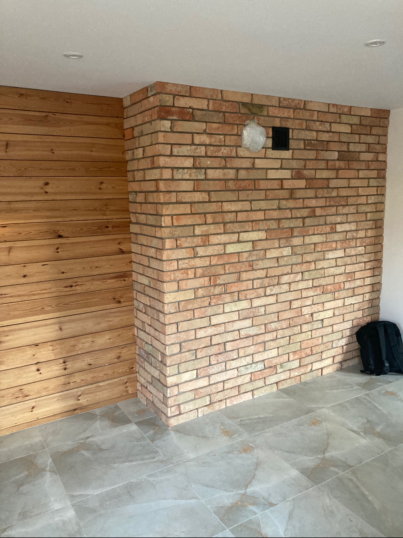 Fragment pokoju z wykończoną ścianą z cegły klinkierowej, drewnianą boazerią i szarą podłogą z płytek imitujących marmur. W tle widoczny czarny plecak.