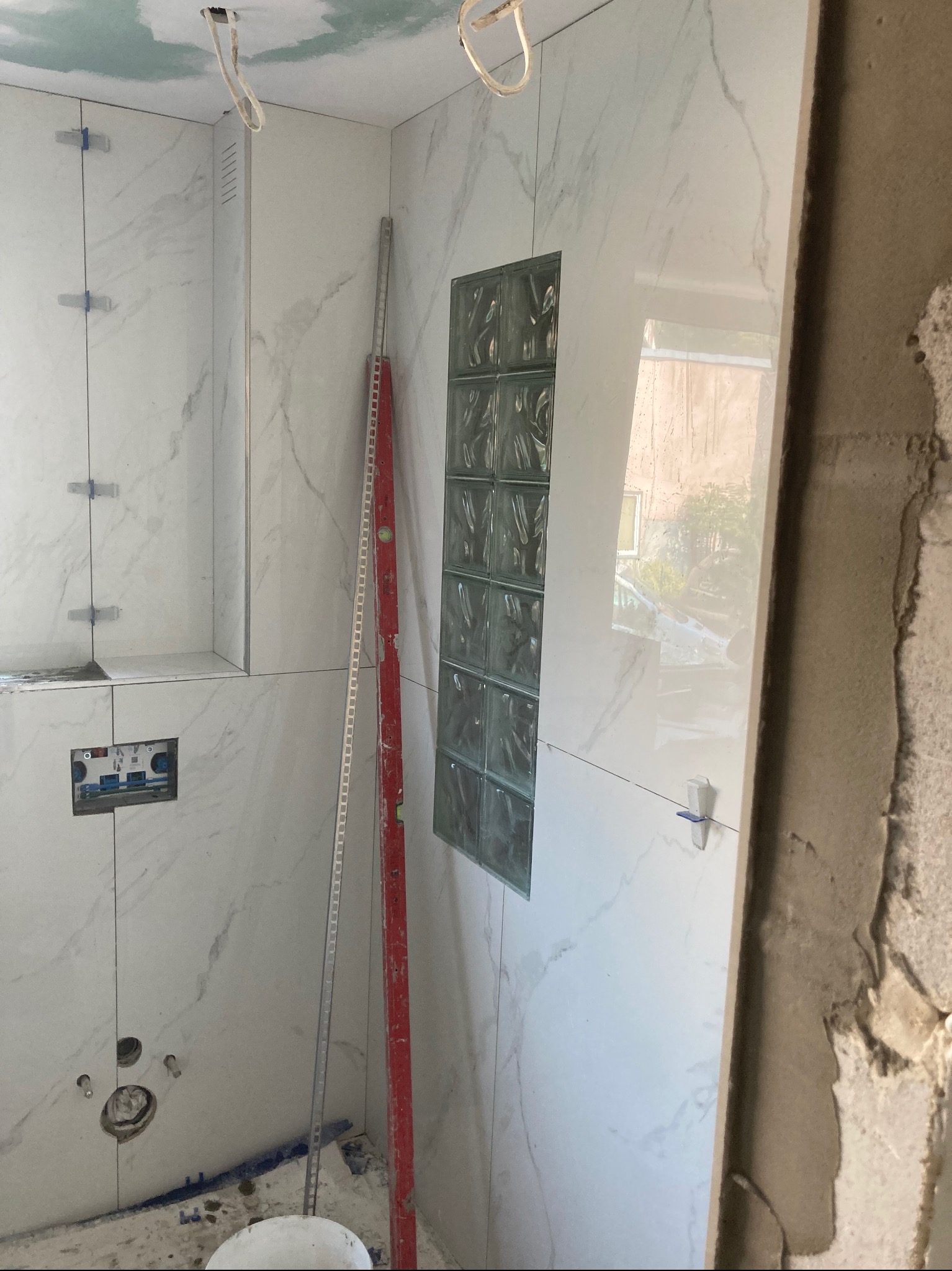 Łazienka w trakcie prac wykończeniowych: białe płytki imitujące marmur z szarymi żyłami, szklane luksfery w ścianie, poziomica oparta o ścianę, widoczne otwory pod armaturę, plastikowe krzyżyki...