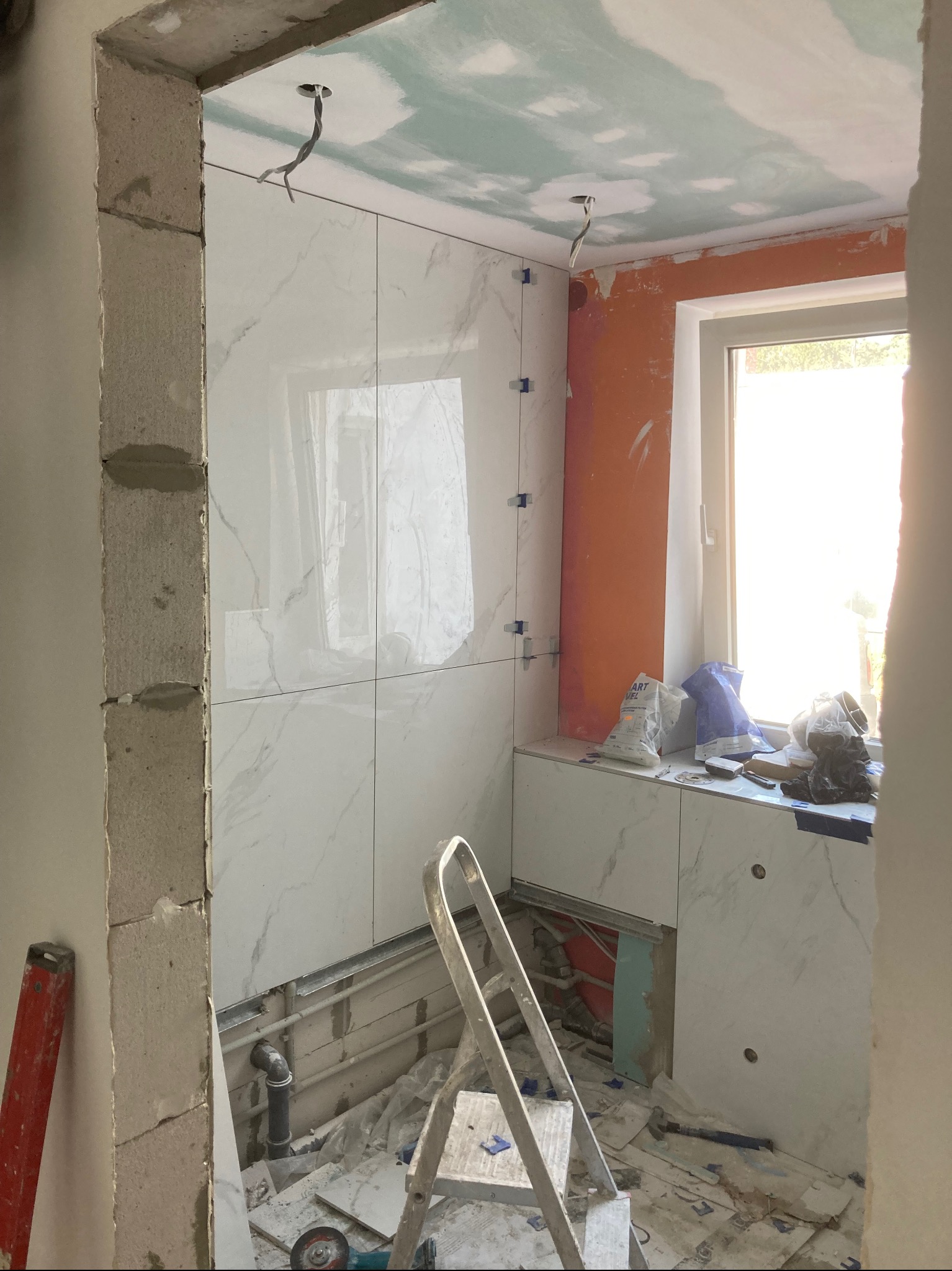 Łazienka w trakcie kładzenia płytek imitujących marmur na ścianach, widoczne krzyżyki dystansowe, drabina, instalacja hydrauliczna i elektryczna, surowy stan wykończenia ścian i sufitu.