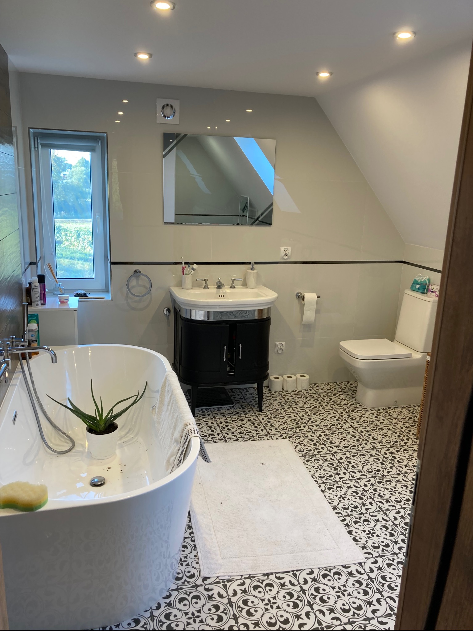 Ukończona łazienka na poddaszu z białą wolnostojącą wanną, czarną szafką pod umywalką, toaletą i wzorzystą czarno-białą podłogą.