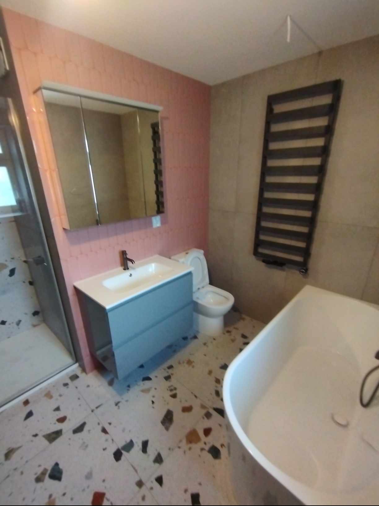 Nowoczesna łazienka z różową ścianą z heksagonalnymi płytkami, szarą szafką pod umywalką, białą toaletą i wanną, oraz grzejnikiem drabinkowym na beżowej ścianie.