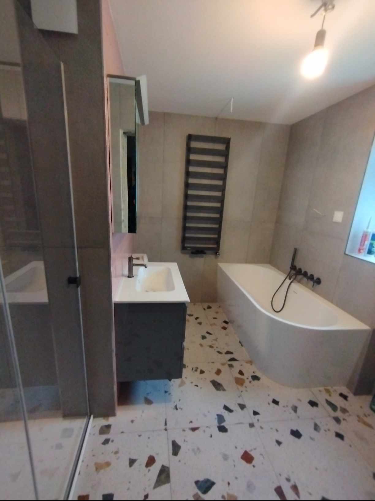 Nowoczesna łazienka z mozaikową podłogą, białą wanną z czarną armaturą, szarą szafką umywalkową i grzejnikiem drabinkowym.