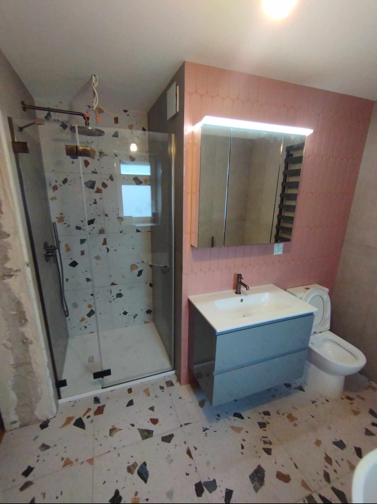 Wykończona łazienka z kabiną prysznicową z czarnymi detalami, umywalką z szarą szafką, toaletą i płytkami w stylu lastryko na podłodze i w kabinie, różowe płytki na ścianie z lustrem z oświetleniem...