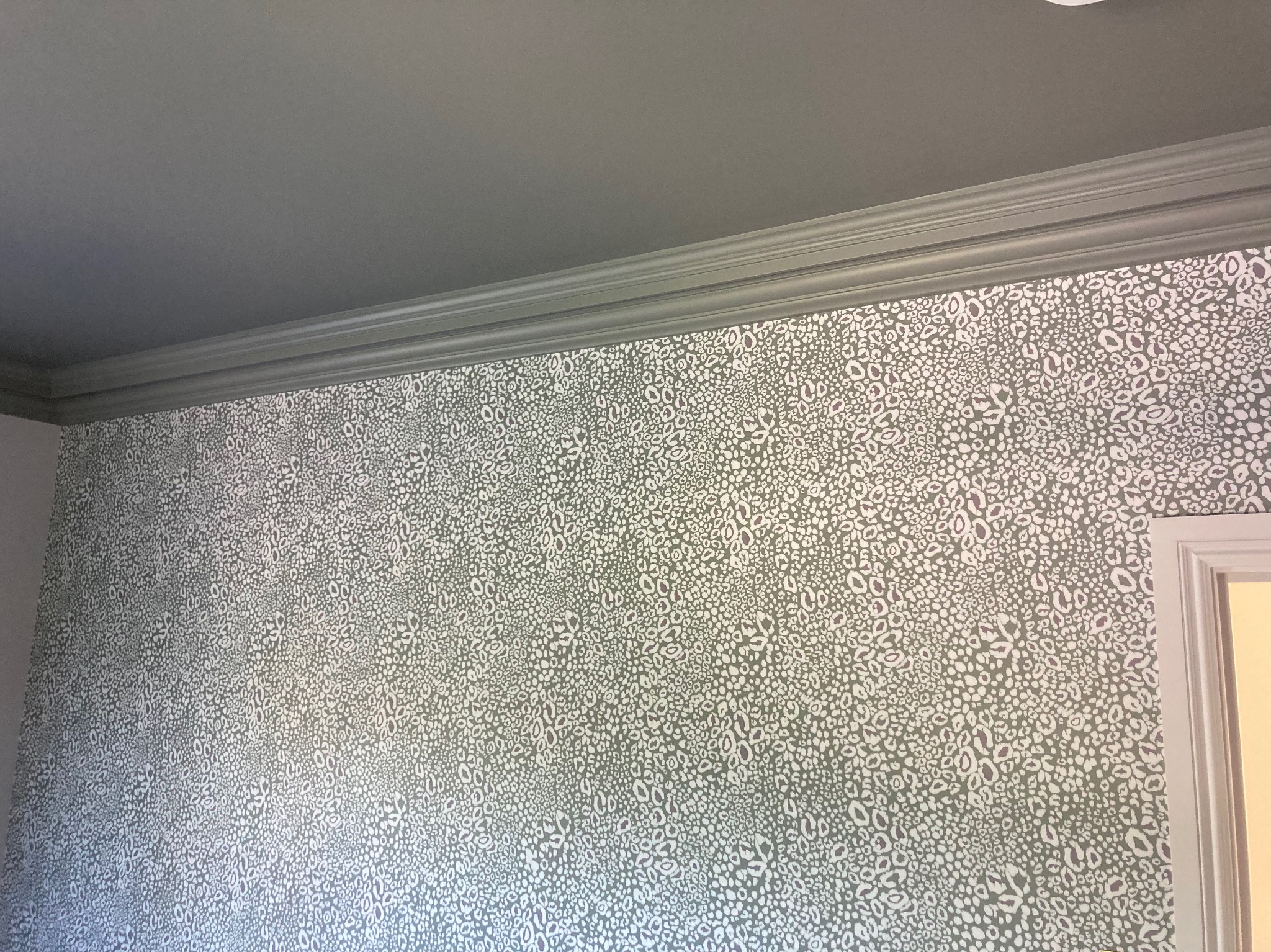 Fragment pokoju z szarą ścianą pokrytą wzorzystą tapetą w odcieniach szarości i bieli, widoczna listwa przysufitowa i fragment futryny drzwi.