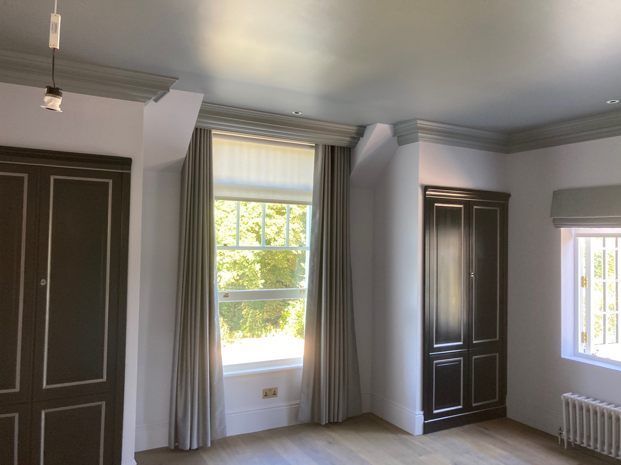 Jasne wnętrze pokoju po remoncie, z dwuskrzydłowymi, ciemnymi szafami wnękowymi z białymi obramowaniami, oknami z kremowymi zasłonami i grzejnikiem żeberkowym pod oknem, wykończone sztukaterią.