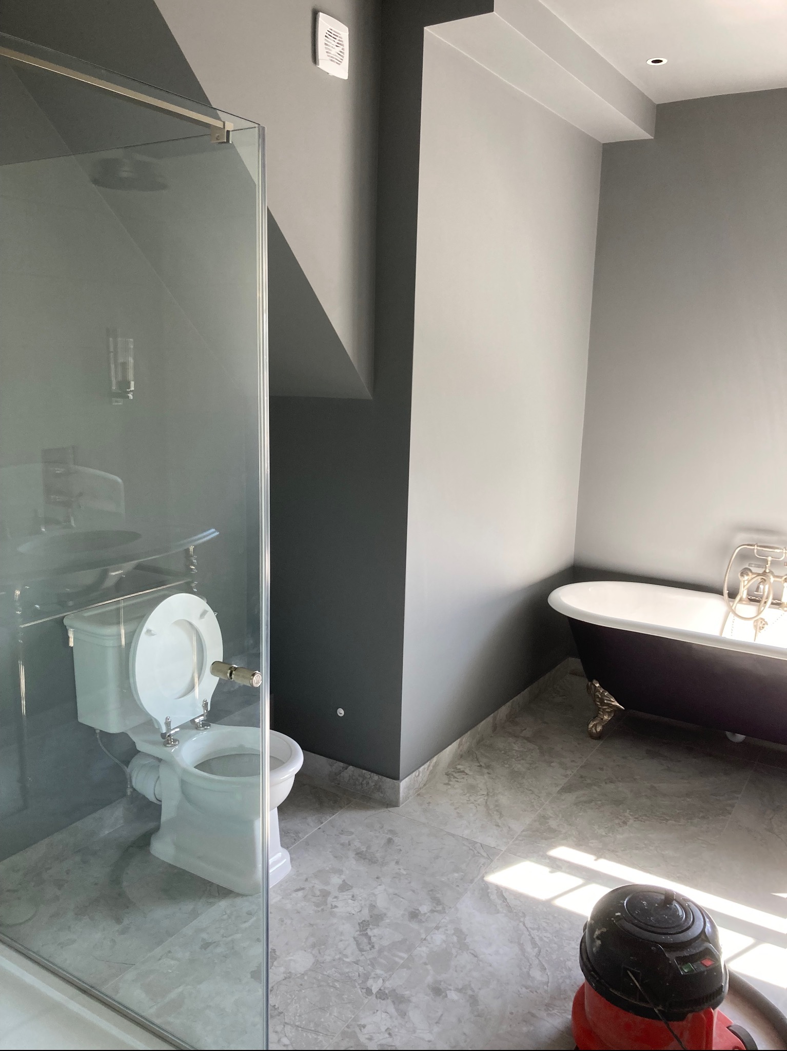 Nowoczesna łazienka w odcieniach szarości z wolnostojącą wanną na ozdobnych nóżkach, toaletą w stylu retro za szklaną kabiną prysznicową i marmurową posadzką, na której stoi odkurzacz.