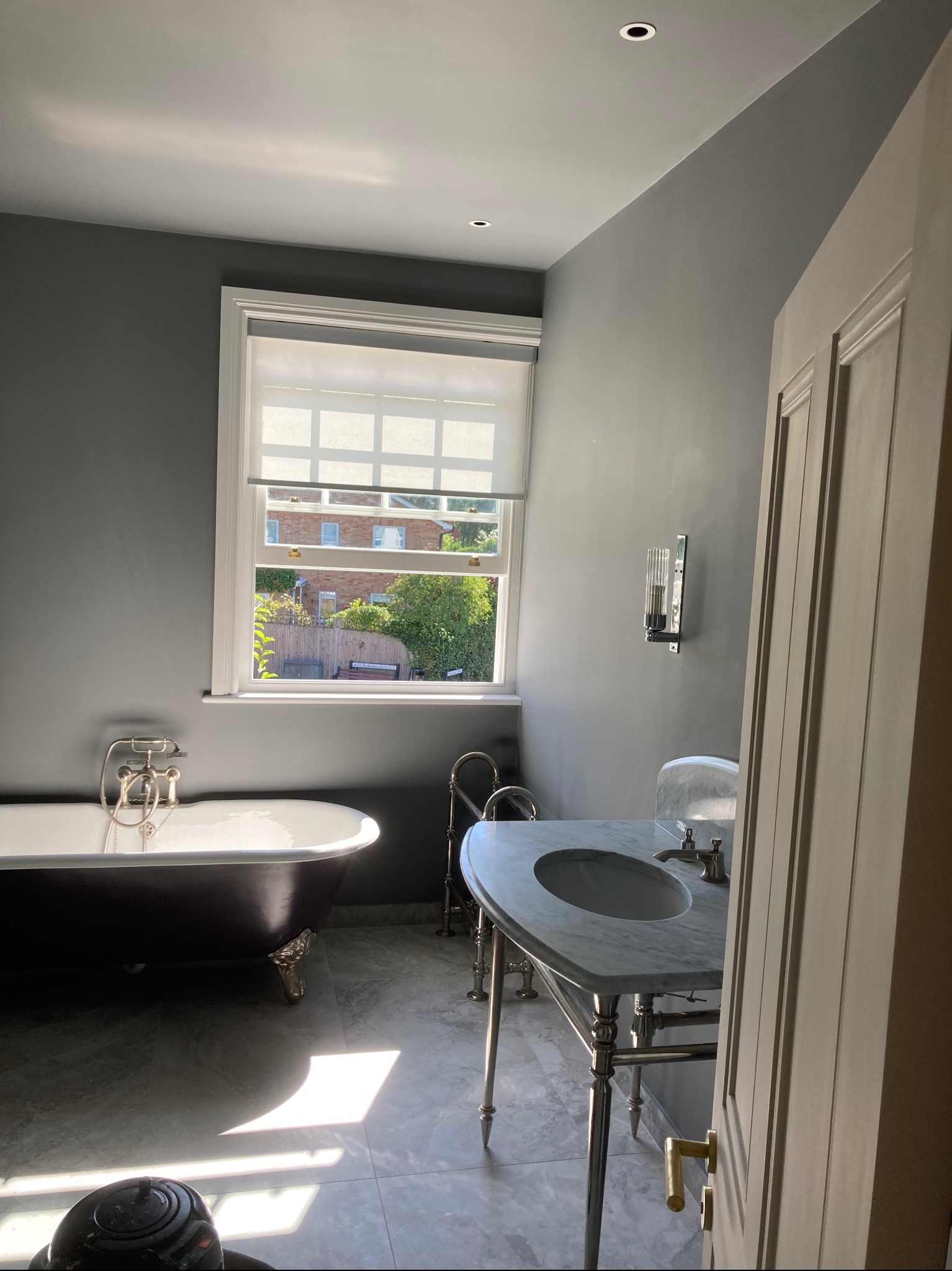 Elegancka łazienka w odcieniach szarości z wolnostojącą wanną na lwich nóżkach, marmurową umywalką na chromowanym stelażu i widokiem na ogród przez okno z roletą.