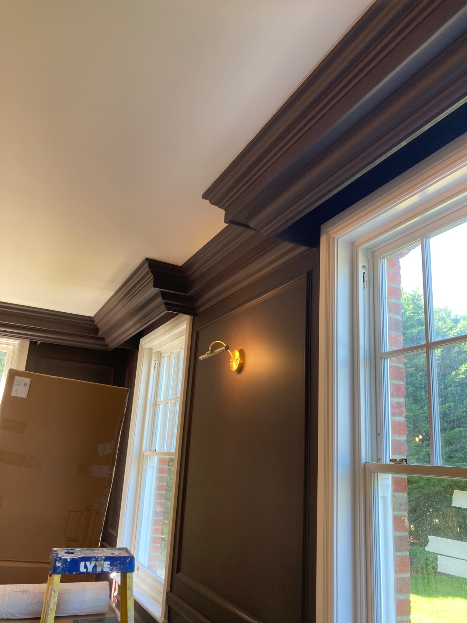 Eleganckie, ciemnobrązowe boazerie i listwy przysufitowe w pokoju z oknami, widoczna drabinka malarska i zabezpieczone okno folią malarską.