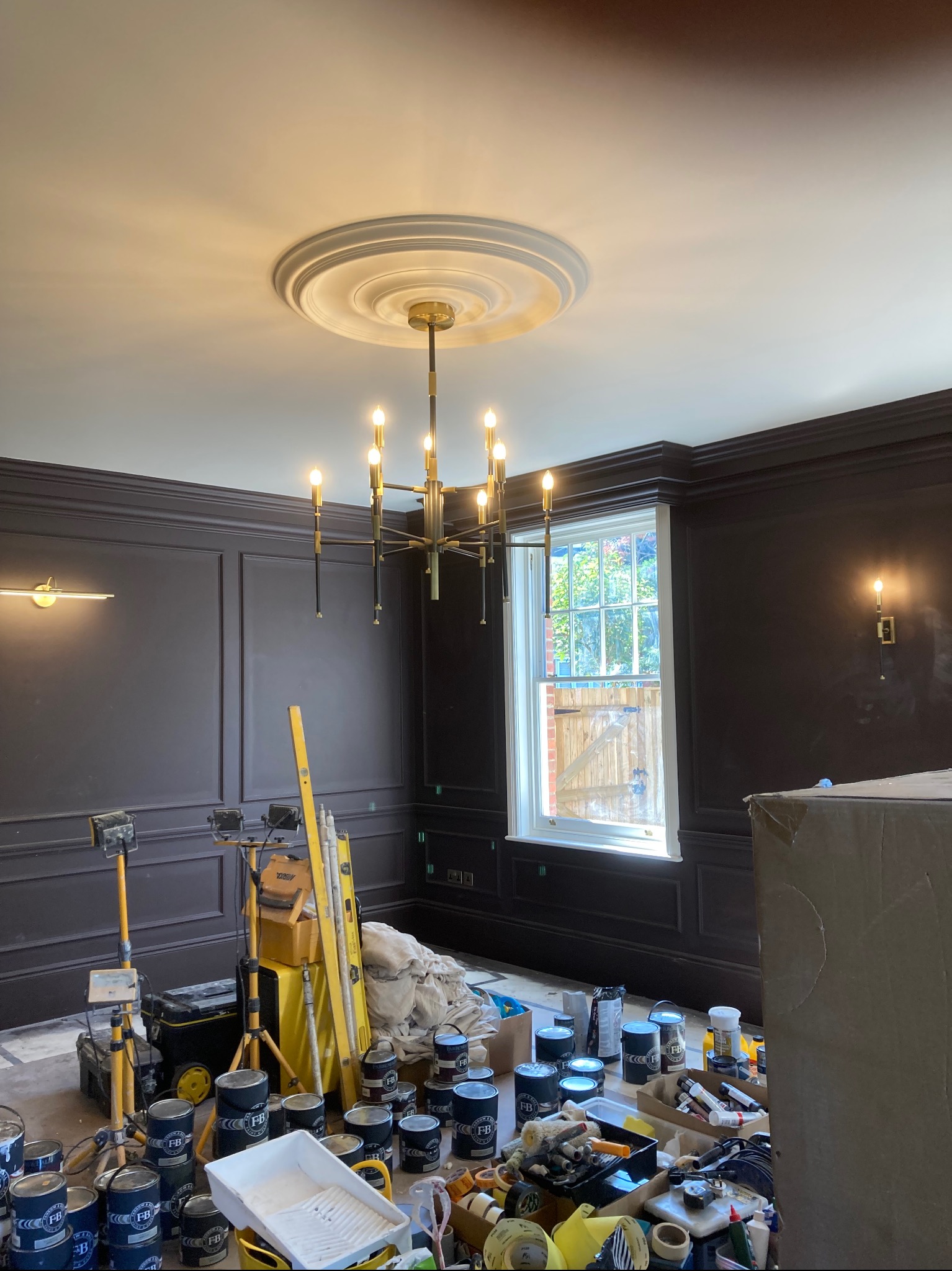 Pokój po malowaniu z ciemnymi ścianami z panelami, widoczne puszki z farbą, drabina i sprzęt oświetleniowy, żyrandol w stylu retro
