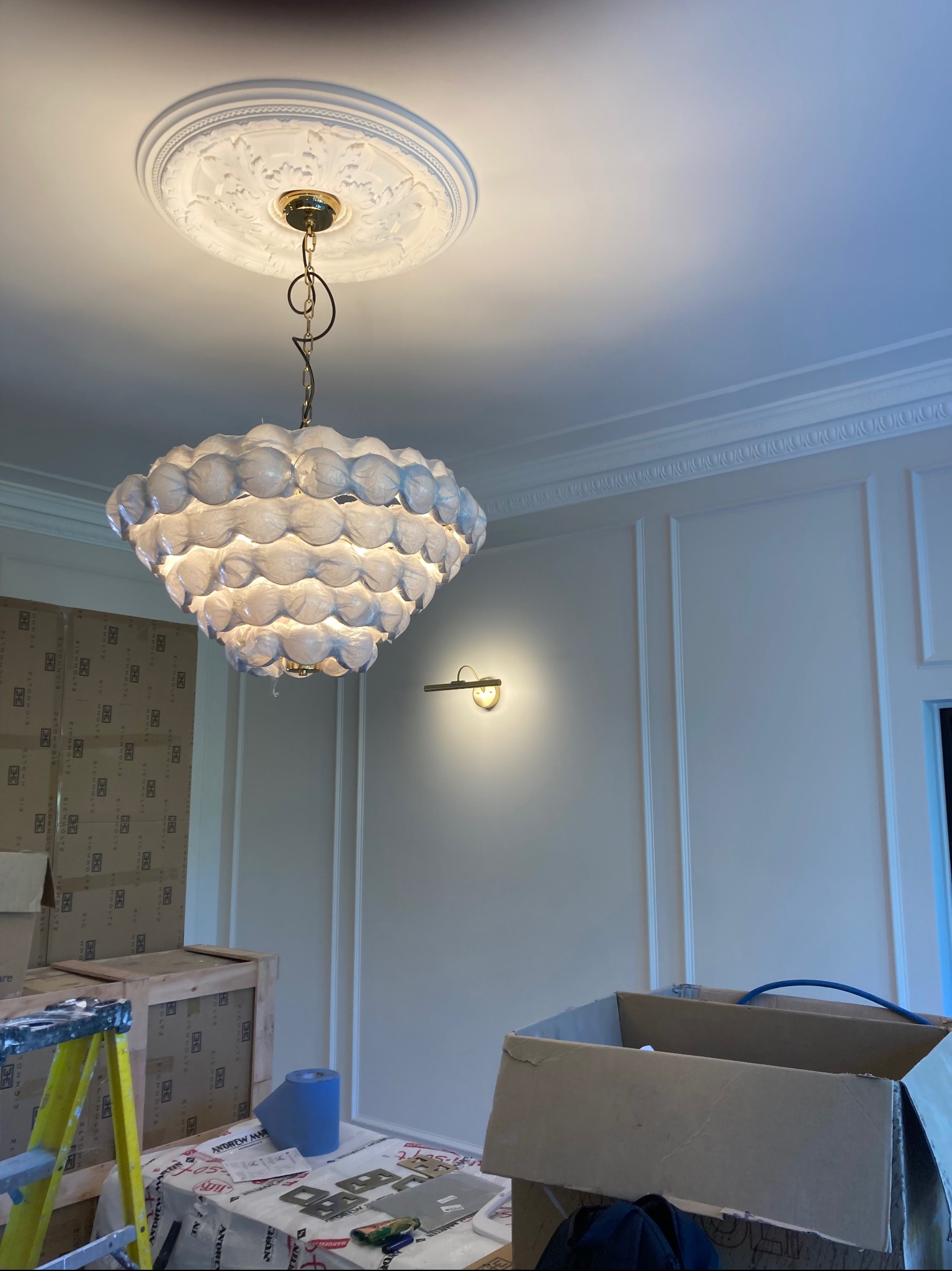 Nowoczesny, kulisty żyrandol z dekoracyjnym rozetą sufitową i listwami ściennymi w trakcie montażu, widoczne kartony i drabina malarska.