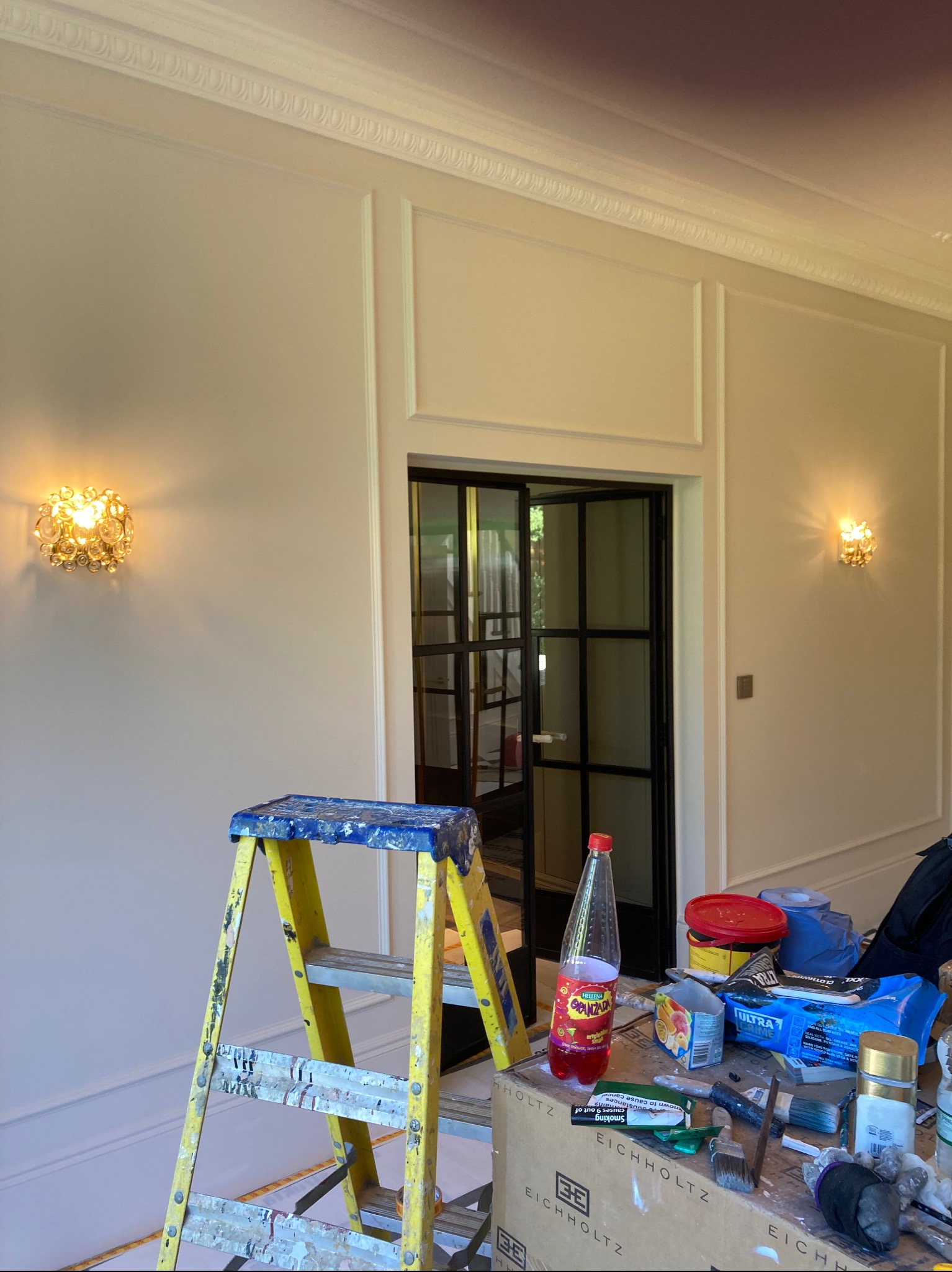 Ściana z dekoracyjnymi panelami i listwami, oświetlona dwoma złotymi kinkietami, widoczny fragment czarnej przeszklonej ścianki działowej, w tle drabina malarska i akcesoria malarskie na kartonie.