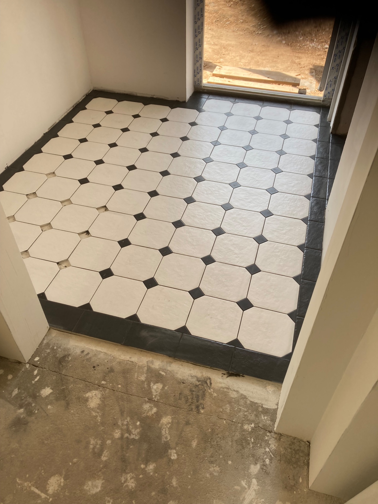 Ułożona podłoga z białych, ośmiokątnych płytek z czarnymi kwadratami na łączeniach, obramowana ciemnymi płytkami, widoczna surowa wylewka podłoża przed fugowaniem, jasne wnętrze z oknem.