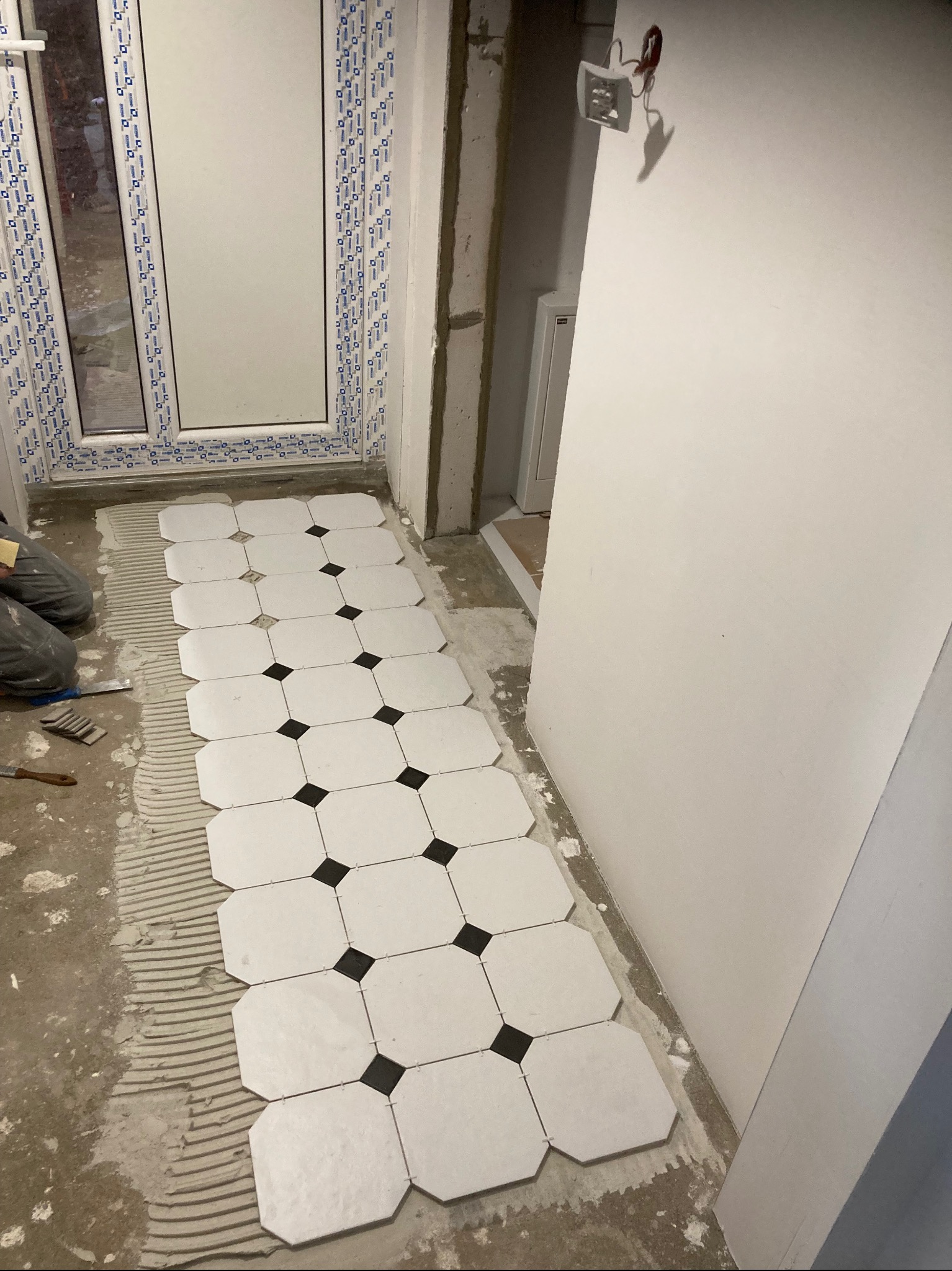 Układanie białych, ośmiokątnych płytek podłogowych z czarnymi wstawkami w pomieszczeniu z oknem PCV, widoczne narzędzia i warstwa kleju.