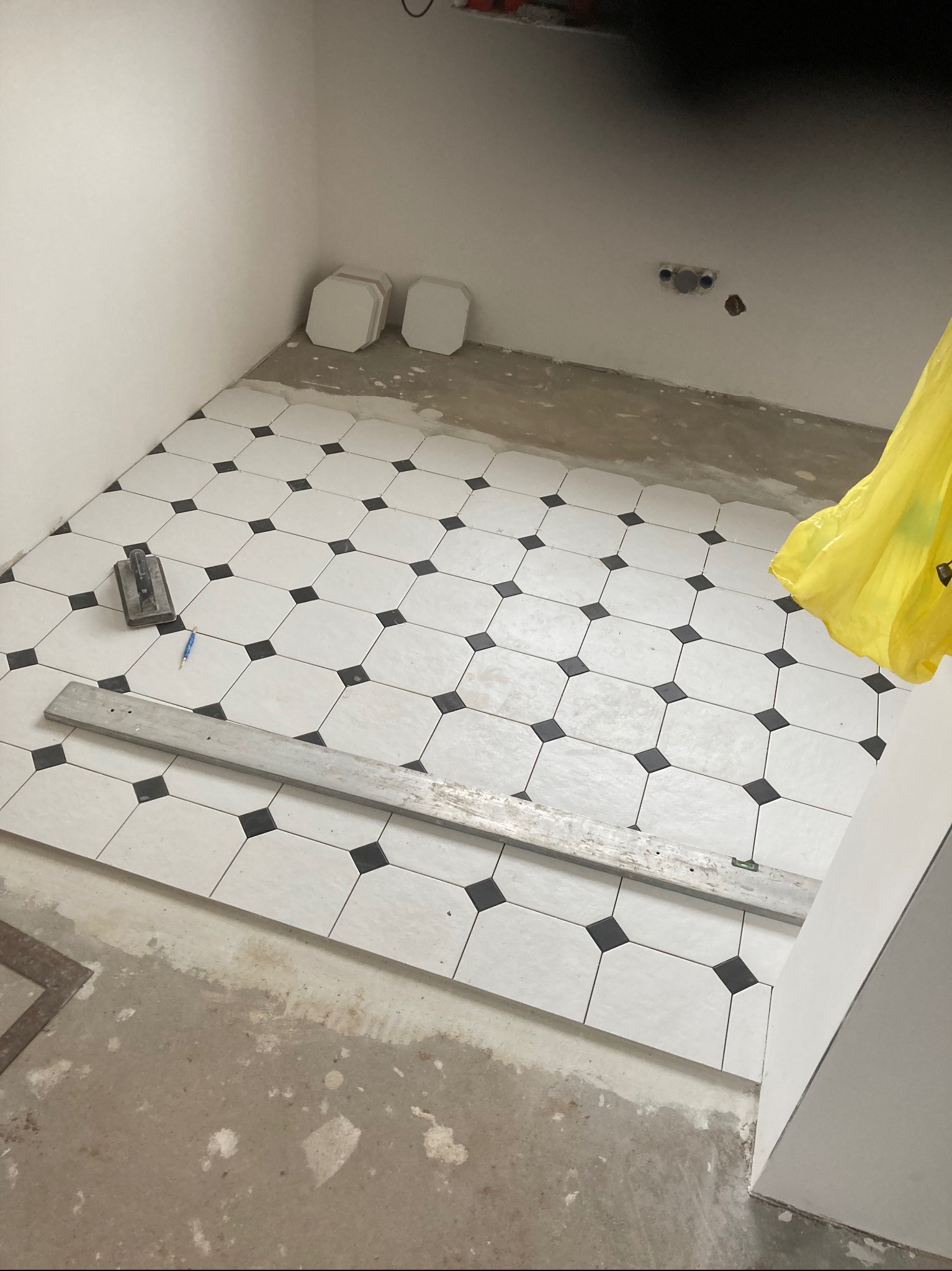 Układanie białych, ośmiokątnych płytek podłogowych z czarnymi kwadratami w narożach, widoczne narzędzia glazurnicze: poziomica, kielnia i ołówek