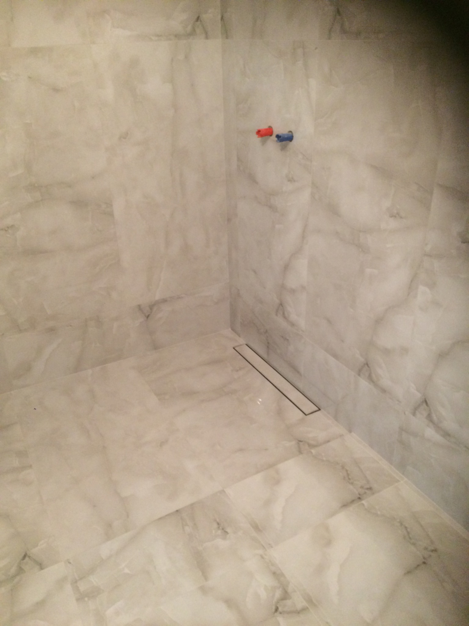 Wykończony brodzik kabiny prysznicowej z odpływem liniowym, wyłożony płytkami imitującymi marmur, widoczne przyłącza ciepłej i zimnej wody.