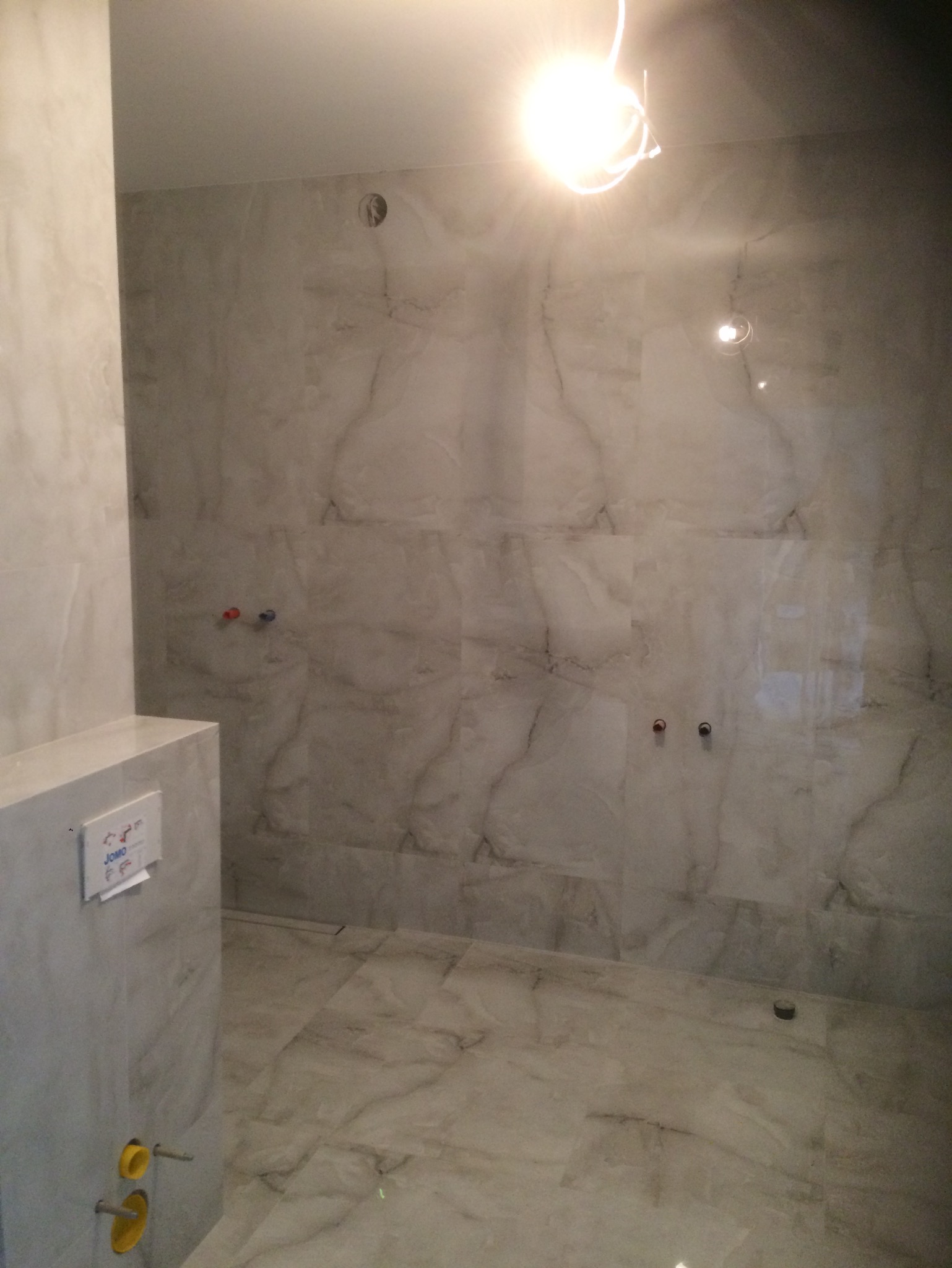 Wykończona łazienka z marmurowymi płytkami na ścianach i podłodze, widoczne przyłącza wodne i element instalacji podtynkowej WC.