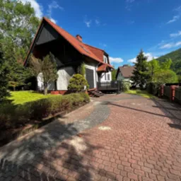 Czerwona kostka brukowa prowadzi do domu z ogrodem. Widok na posesję w słoneczny dzień, z elementami zieleni i górskim krajobrazem w tle.