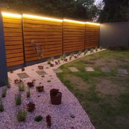 EuNike Architektura Krajobrazu Eunika Wiatr - Nowoczesny ogród po zmroku: podświetlony drewniany płot, ścieżka z kamyczków i płyt, trawnik. Minimalistyczny design z subtelnym oświetleniem.